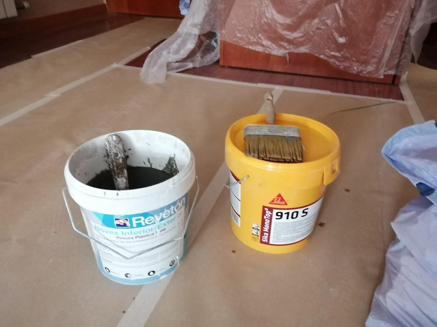 Dos cubos de pintura y una brocha sobre papel protector dentro de una habitación en renovación.