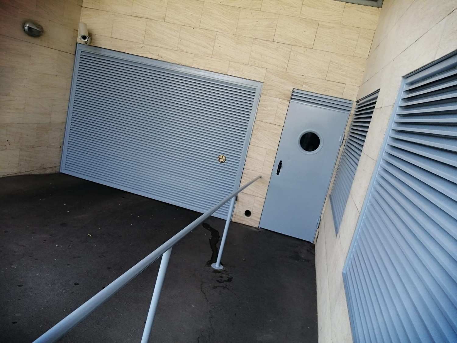 Rampa de acceso con pasamanos metálico junto a un edificio beige y puertas grises cerradas.