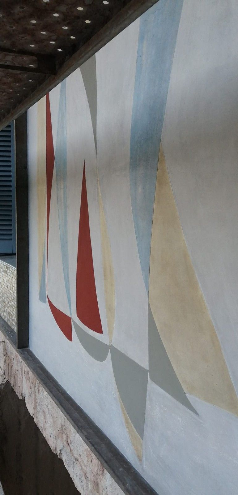 Mural abstracto con formas geométricas rojas, azules y beige que recuerdan a velas sobre una superficie texturizada.