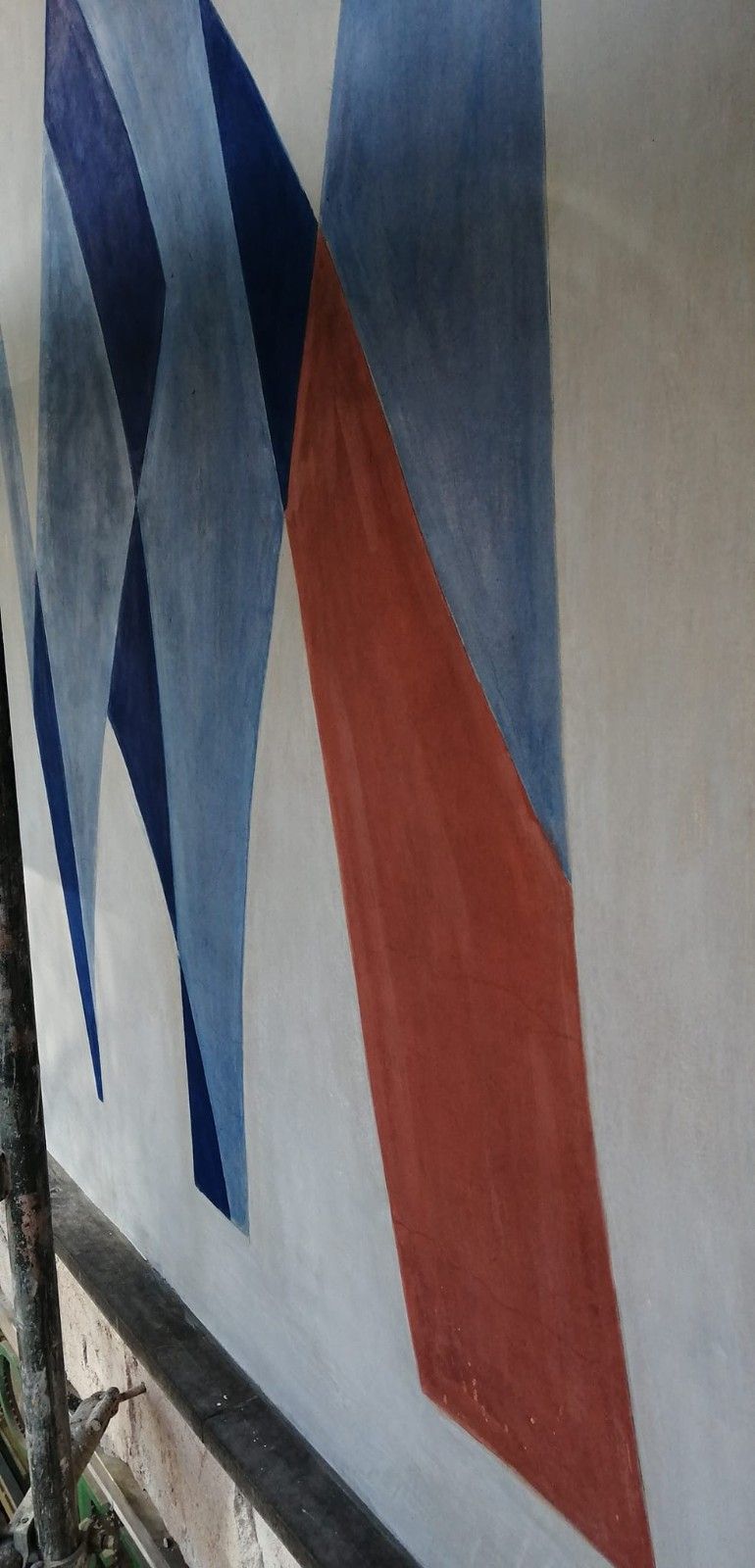 Mural geométrico abstracto con formas angulares azules, blancas y rojas sobre una pared clara.