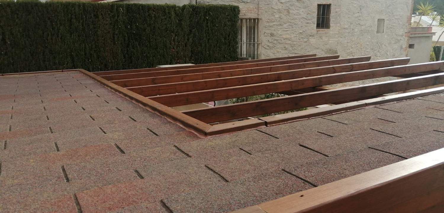Terraza en la azotea con adoquines rojizos y vigas de madera, bordeada por un seto y un muro de piedra.