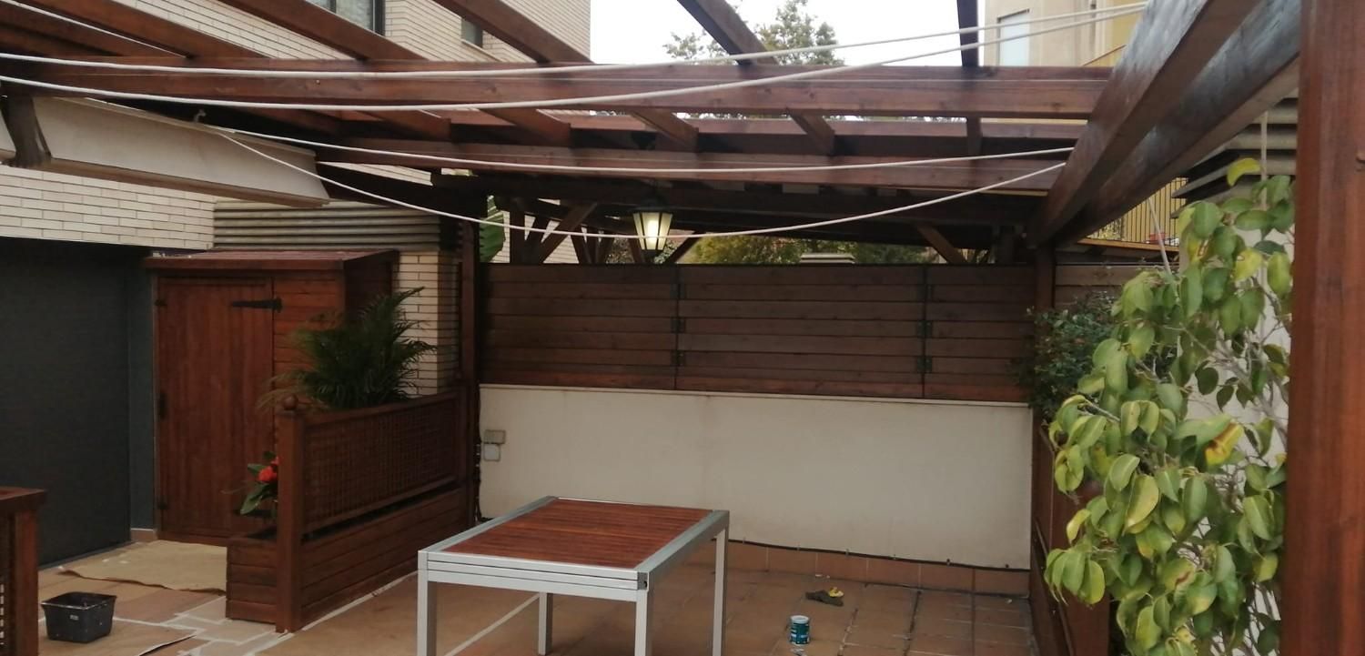 Patio cubierto con vigas de madera, una mesa y plantas en macetas junto a una pared de ladrillo.