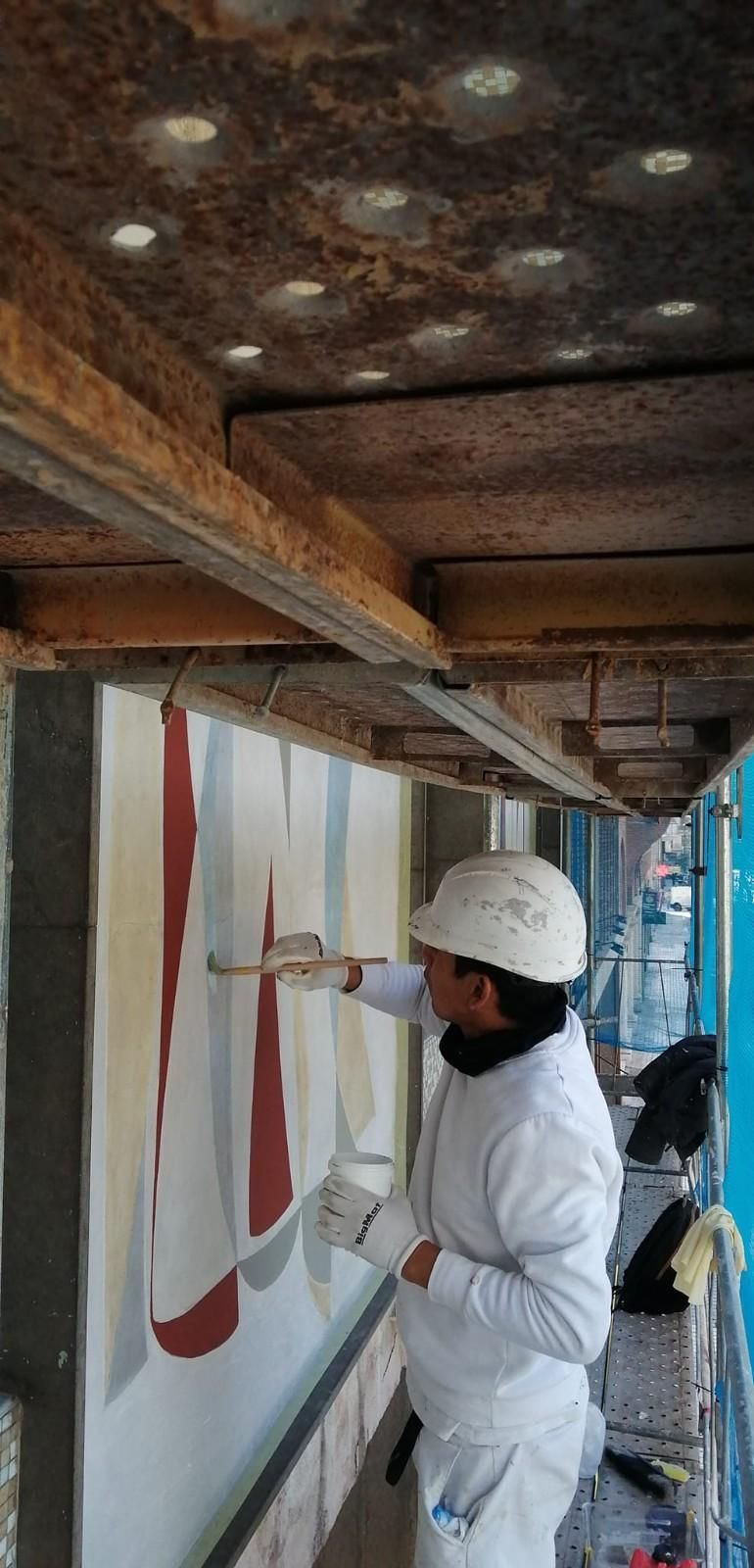 Un trabajador con equipo de seguridad blanco inspecciona una pared de andamios con formas rojas pintadas y un techo dañado.