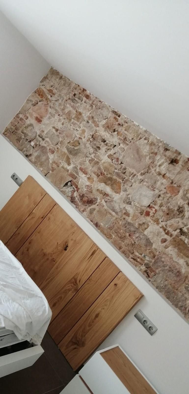 Vista inclinada del dormitorio con pared decorativa de piedra, cabecero de madera y esquina de la cama.