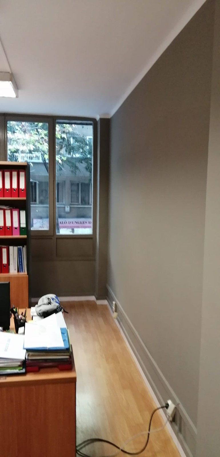 Pasillo de oficina estrecho con un escritorio de madera, estanterías, ventana y cable eléctrico a lo largo del suelo.