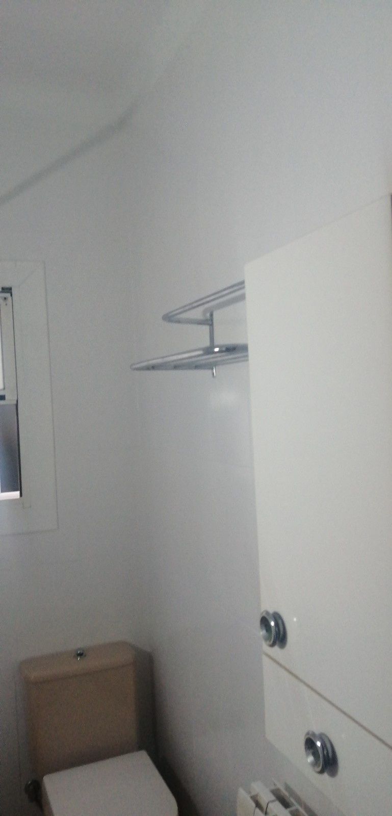 Baño pequeño con paredes blancas, un estante para toallas, una ventana y una caja de cartón en el suelo.