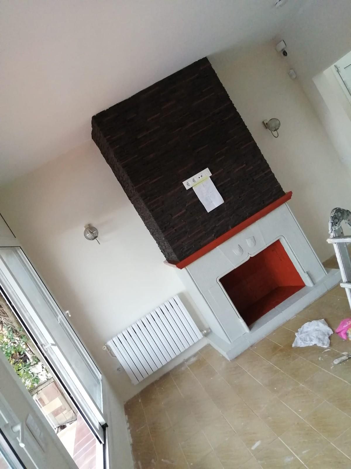 Vista oblicua de una luminosa sala de estar con suelo de baldosas beige, chimenea blanca y pared decorativa oscura.