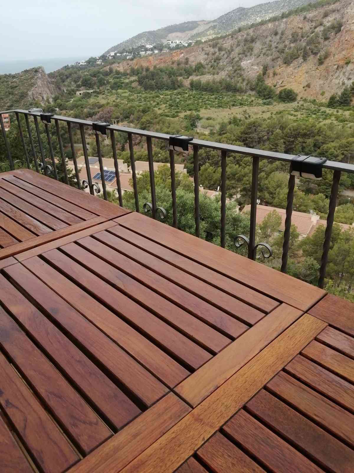 Terraza de madera con barandilla negra con vistas a un valle verde y montañas rocosas bajo un cielo nublado.