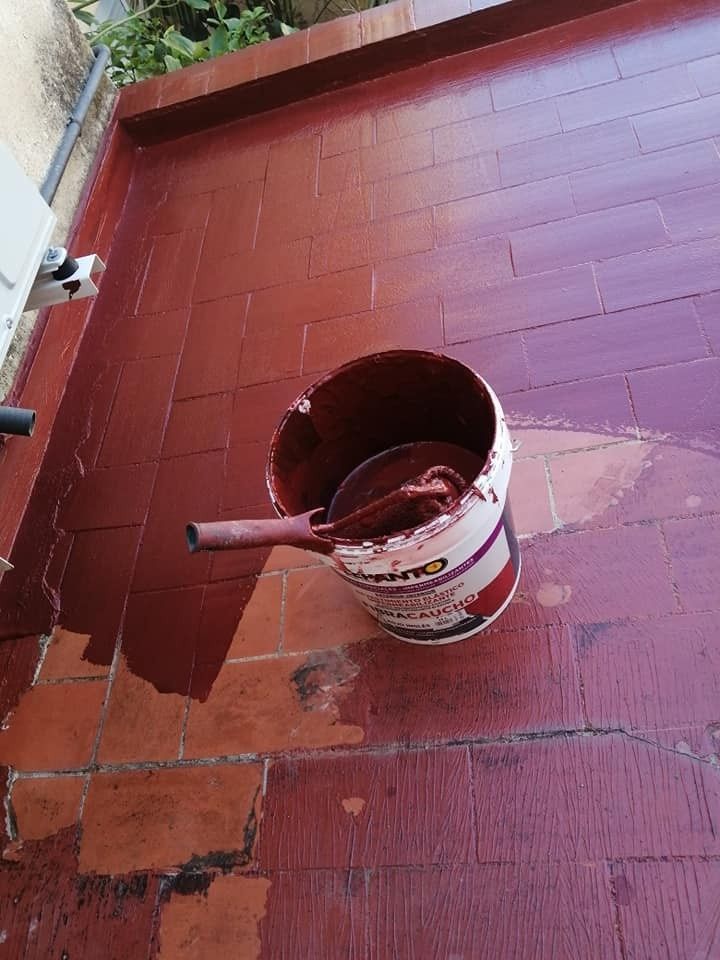 Cubo de pintura con pintura roja y pincel sobre un suelo de baldosas rojas cerca de una pared y una barandilla.