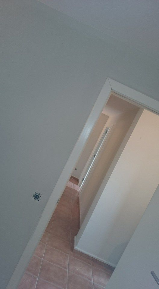 Vista inclinada de un pasillo con paredes blancas, suelo de baldosas beige y una puerta abierta que da a una habitación.