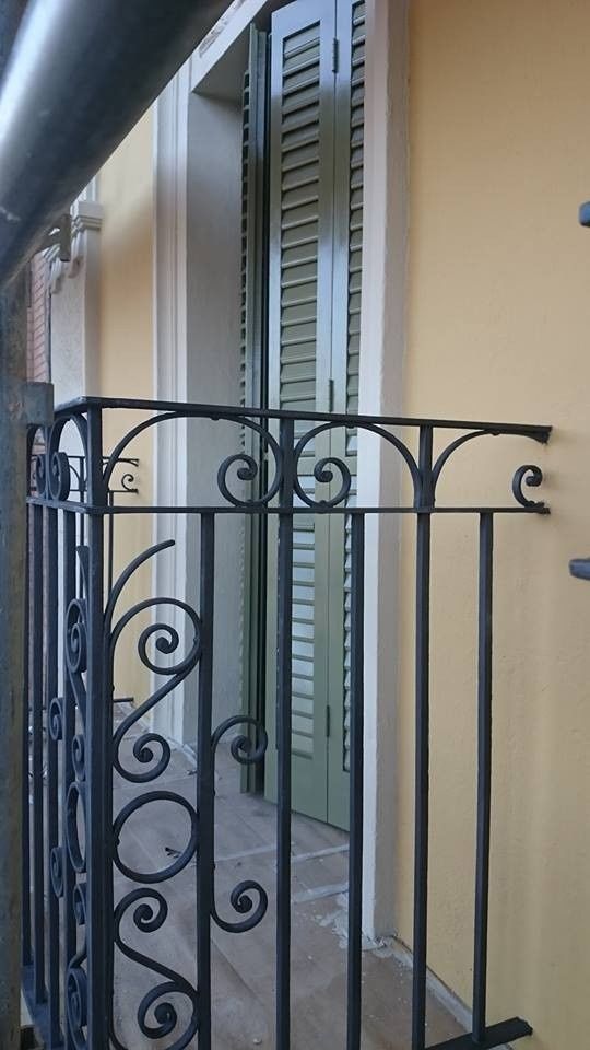 Balcón estrecho con barandilla negra ornamentada, paredes beige y una puerta y ventana con contraventanas verdes detrás.