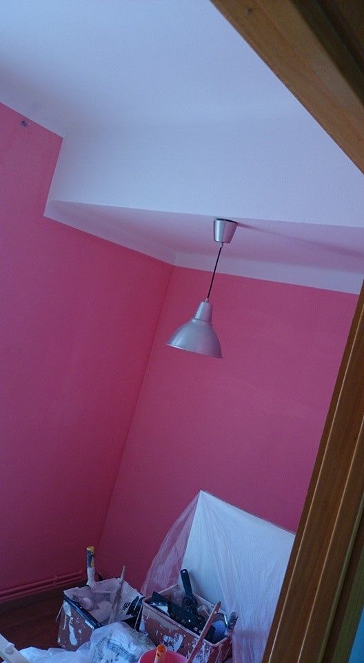 Rincón de habitación rosa con molduras blancas en el techo, lámpara colgante y escritorio abarrotado de objetos.