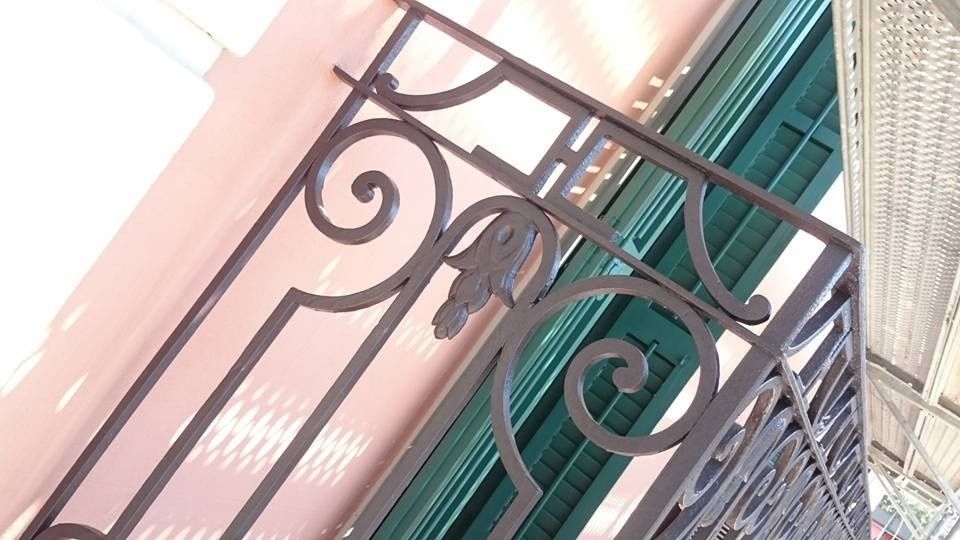 Barandilla decorativa de hierro forjado en un balcón, junto a contraventanas verdes, en un edificio de color melocotón.