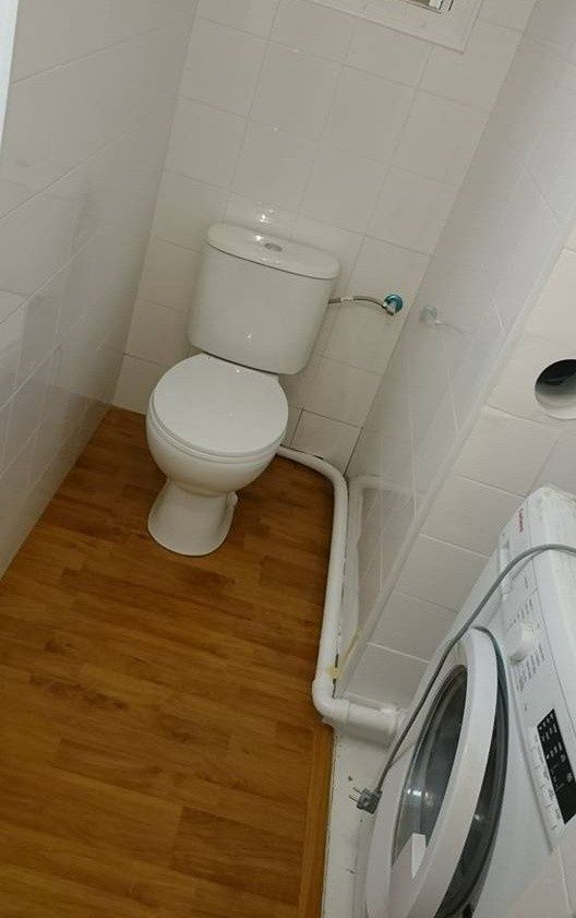 Baño pequeño con inodoro blanco, paredes alicatadas, suelo de madera y lavadora a la derecha.