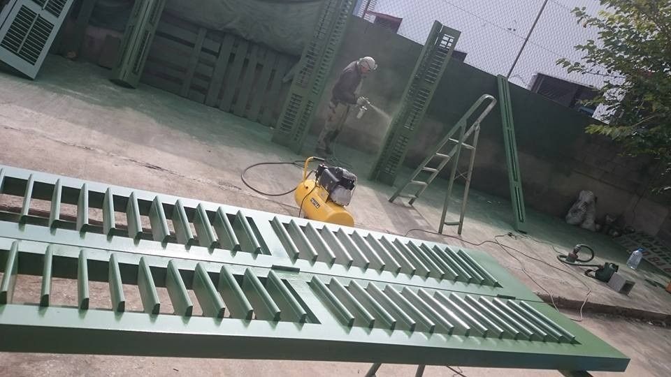 Persianas metálicas verdes extendidas en el suelo de un taller, con una herramienta eléctrica amarilla y una escalera cerca.