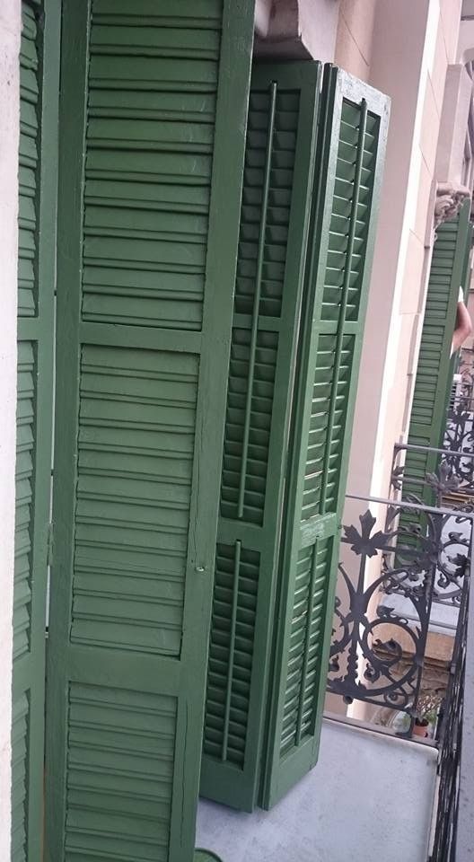 Persianas verdes de lamas en el balcón de un edificio, junto a una barandilla de hierro forjado y la pared contigua.