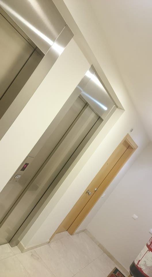 Vista inclinada de un vestíbulo de ascensor con puertas de acero inoxidable y madera sobre una pared beige de un pasillo.