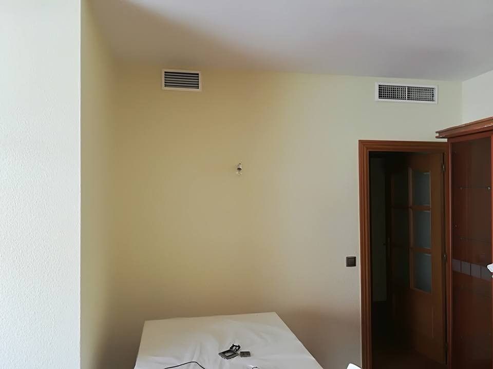 Habitación vacía de color beige con una cama, rejillas de ventilación en la pared y una puerta de madera abierta.