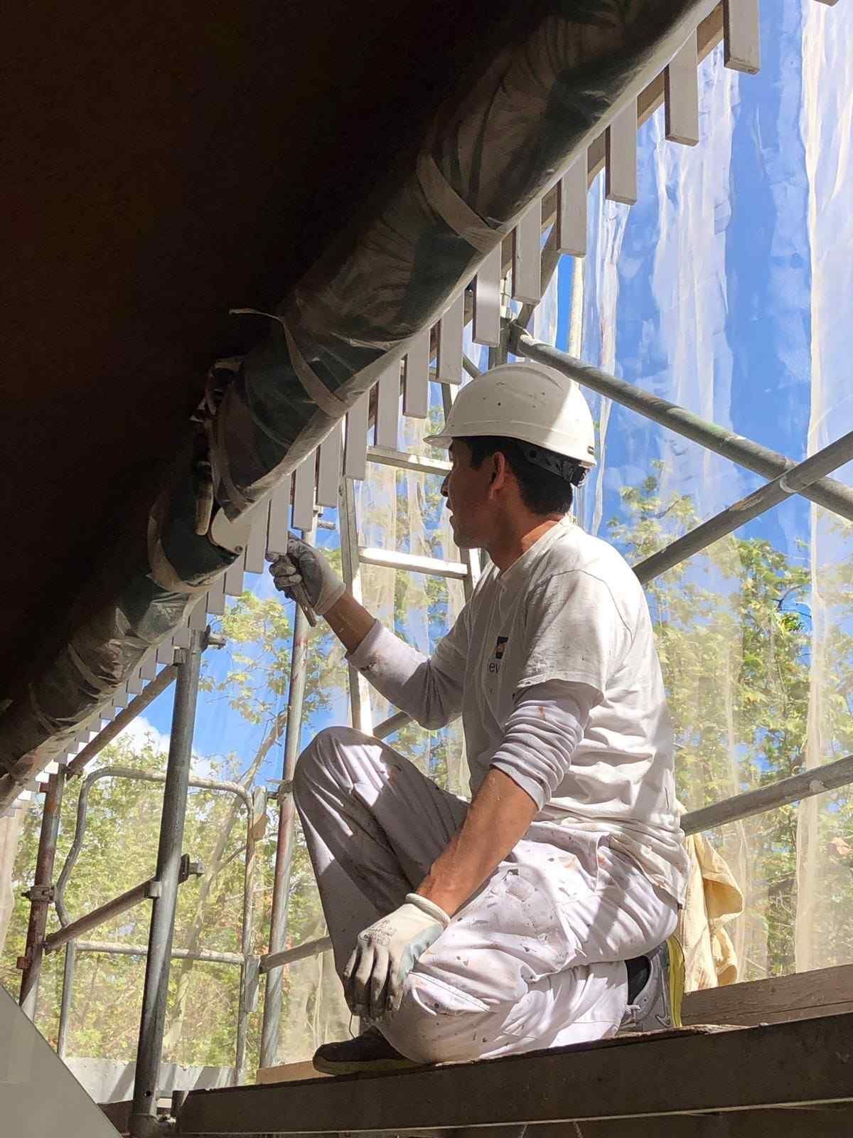 Un trabajador vestido de blanco pinta una estructura sobre un andamio, cubierta con una lona azul.