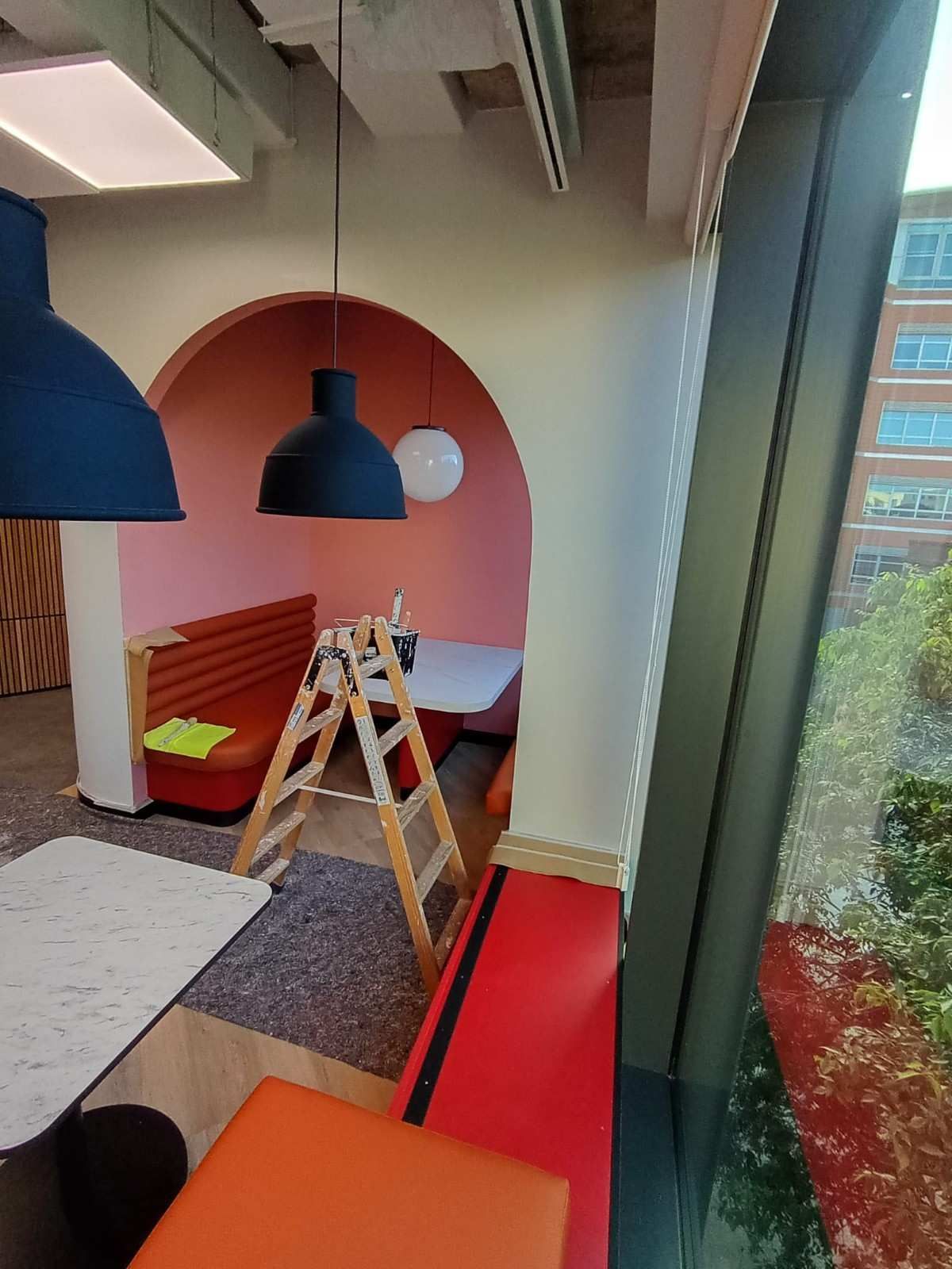 Sala de estar de oficina moderna con rincón de asientos arqueado rojo, lámparas azules colgantes, escalera y gran ventanal.