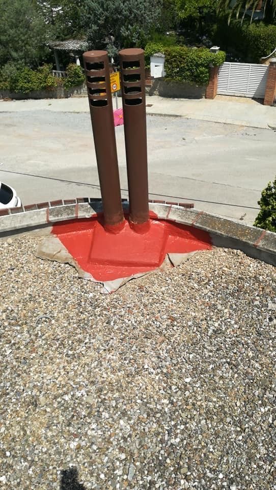 Dos tuberías metálicas de color óxido sobre una base roja en un patio de grava, junto a una acera de hormigón.