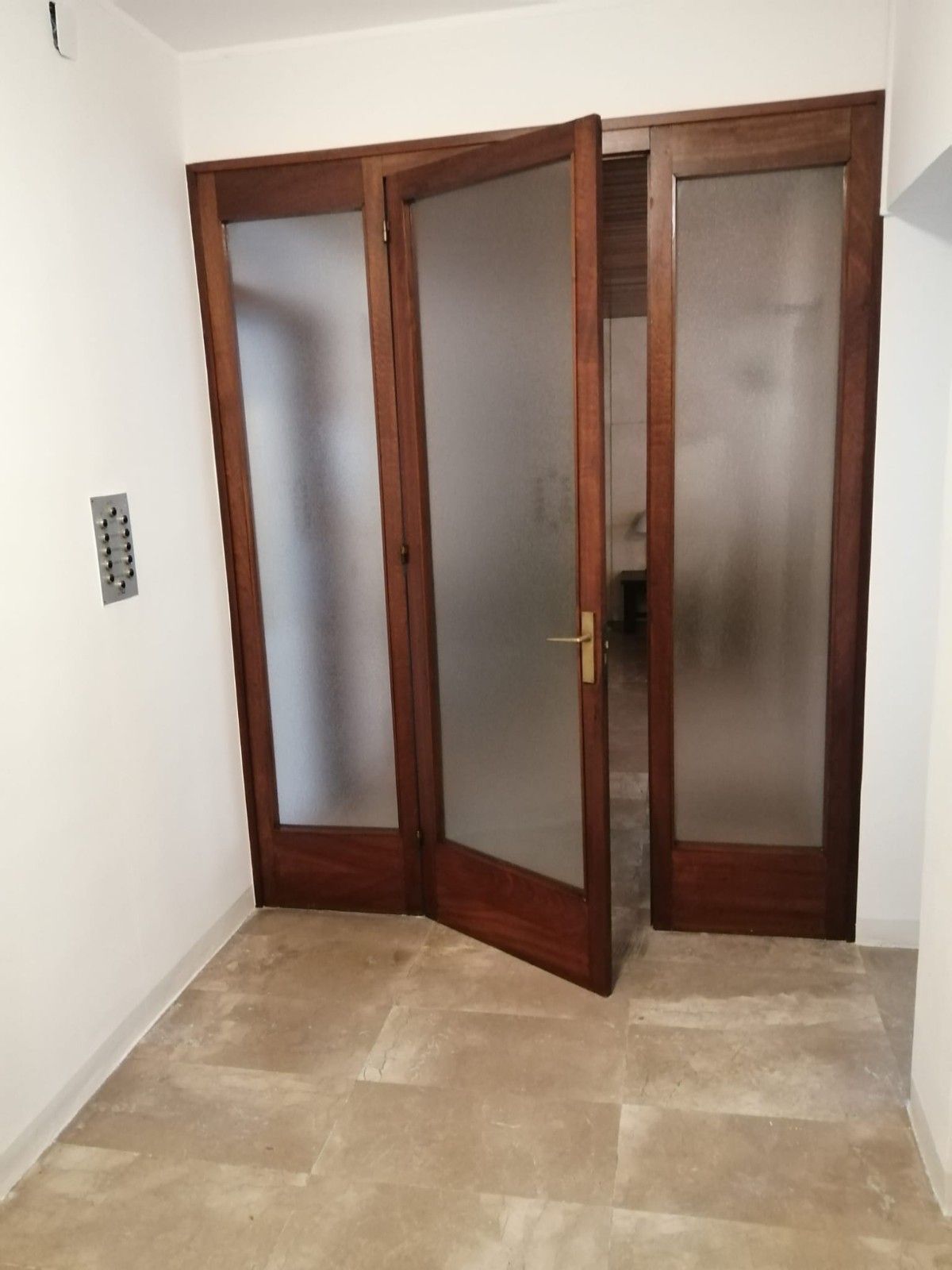 Puertas de madera y cristal parcialmente abiertas en un pasillo vacío con suelo de baldosas beige.