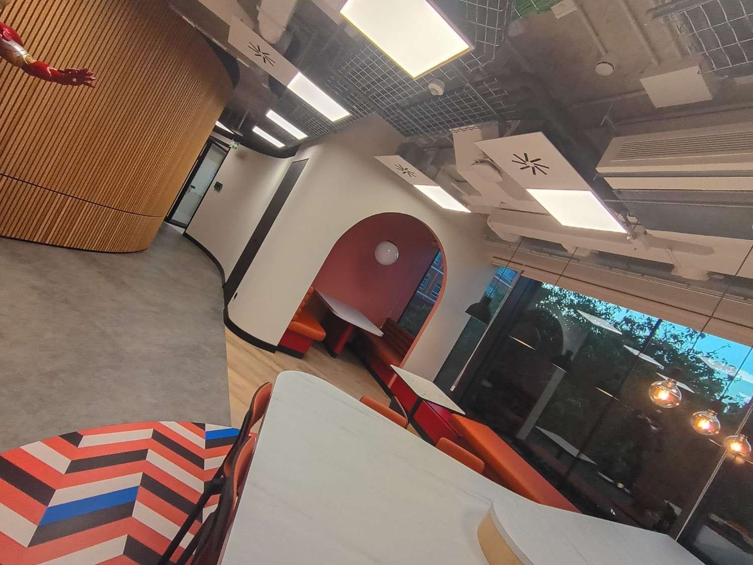 Interior de cafetería moderna con cabinas rojas, sillas estampadas, grandes ventanales y luces en el techo.