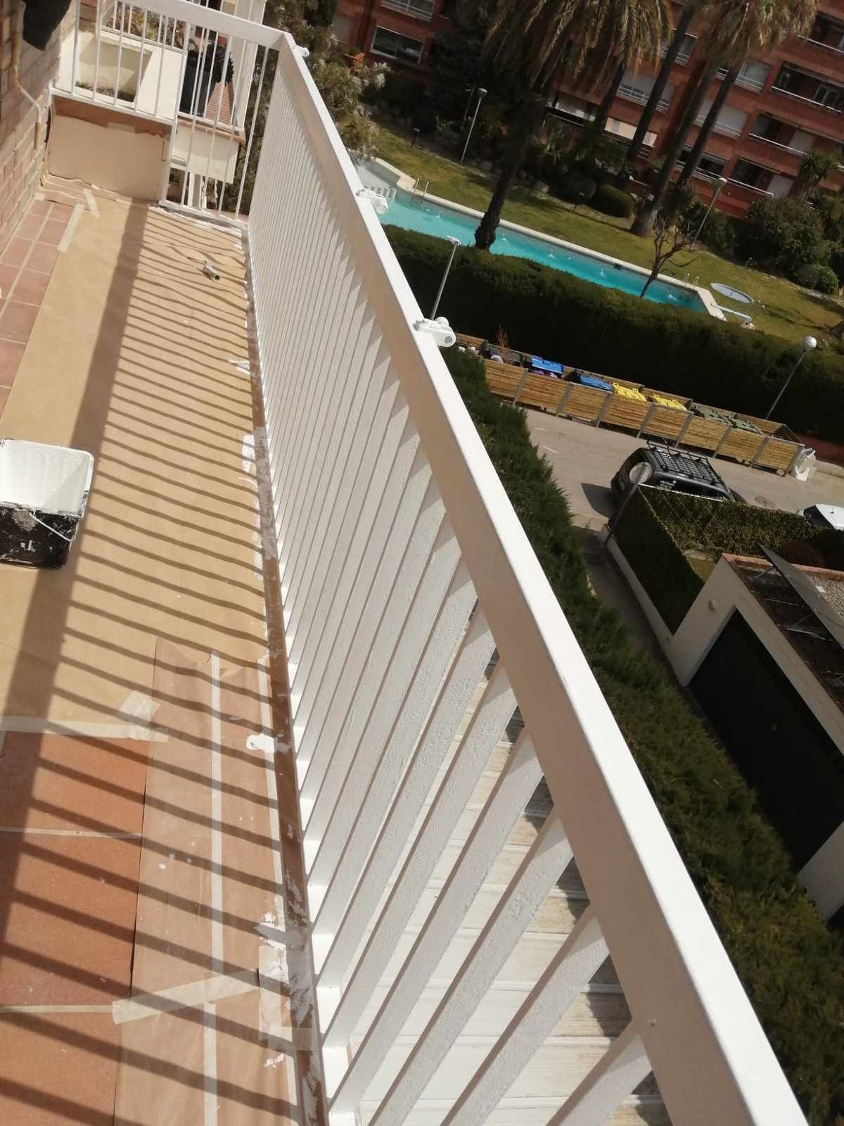 Pasillo con balcón, barandilla blanca y baldosas color canela, con vistas a una piscina y al patio del edificio.