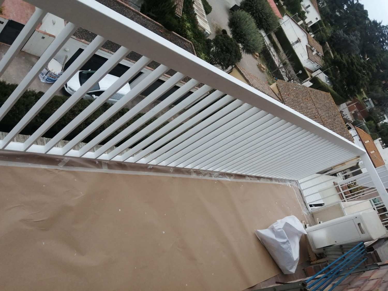 Balcón con barandilla blanca, suelo cubierto y una pequeña parrilla junto a macetas y vistas a los tejados del vecindario.
