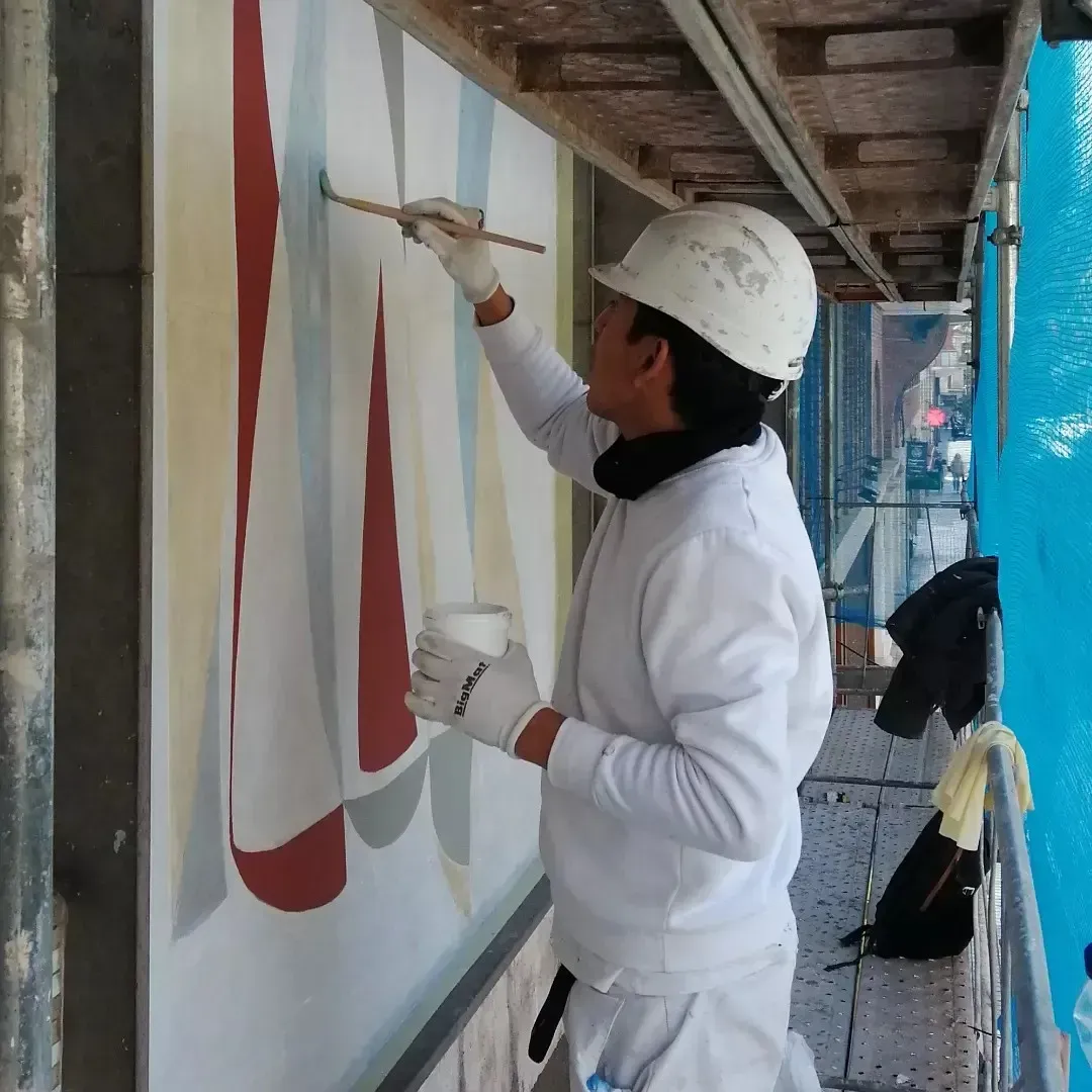 Pintor con traje blanco aplicando pintura a un mural en una pasarela exterior sostenida por andamios.