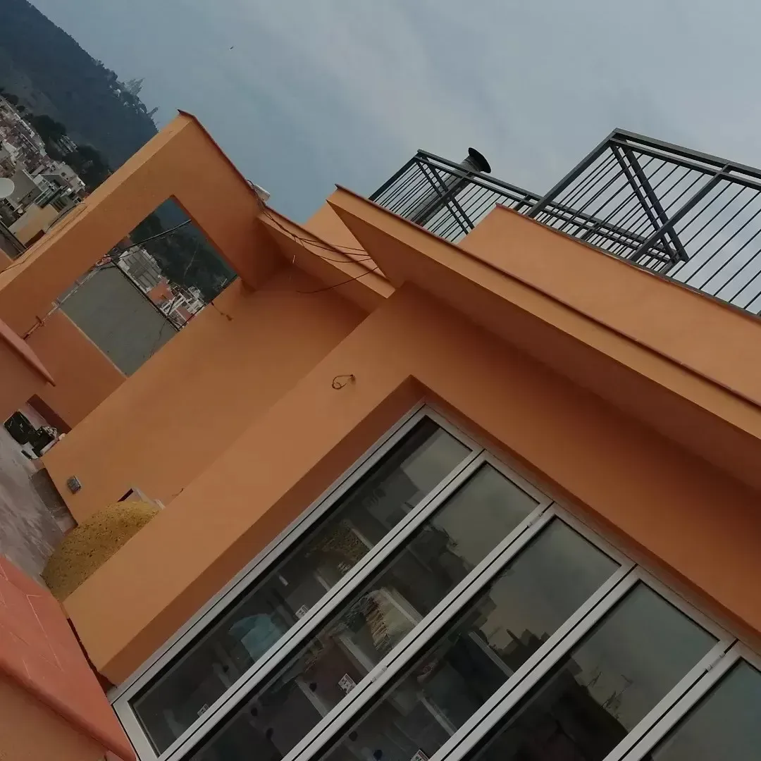 Vista inclinada de una casa naranja con grandes ventanales y una barandilla en el tejado contra un cielo nublado.