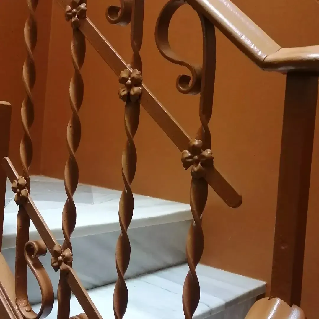 Barandilla de escalera de metal retorcido junto a escalones de mármol blanco sobre una pared de color naranja cálido.