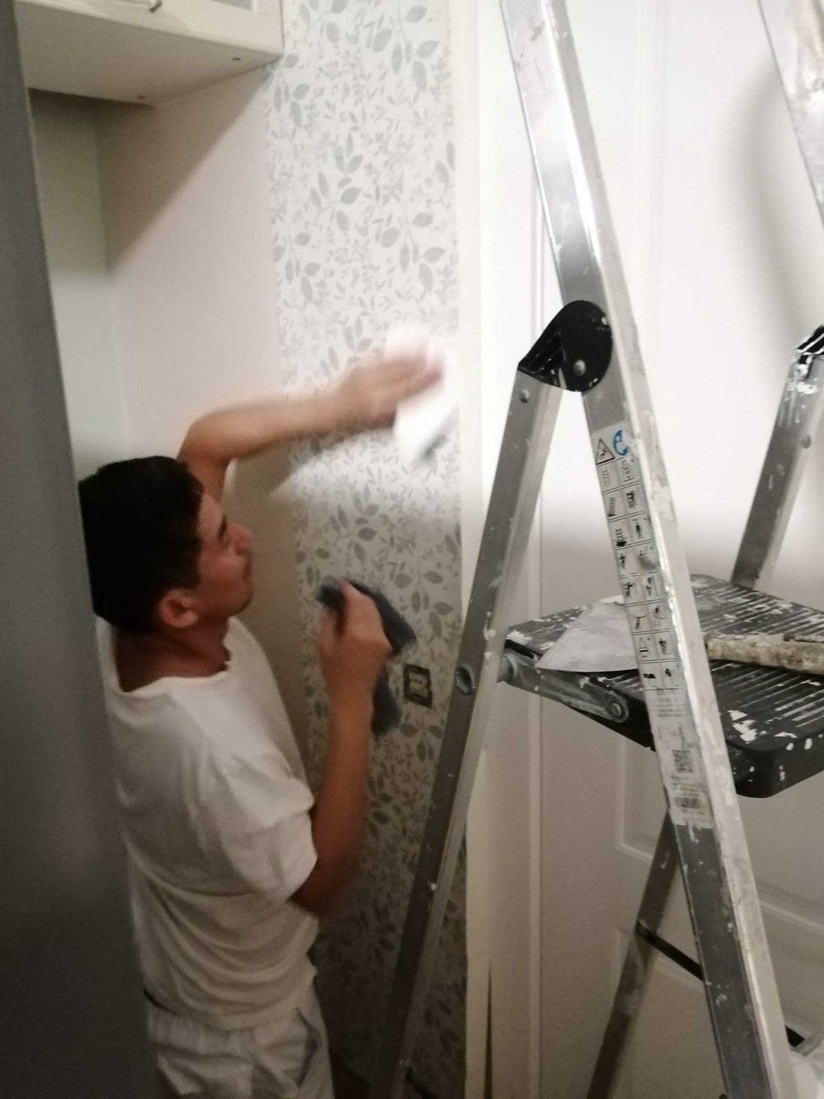 Persona usando una espátula para quitar papel tapiz junto a una escalera en una habitación estrecha.