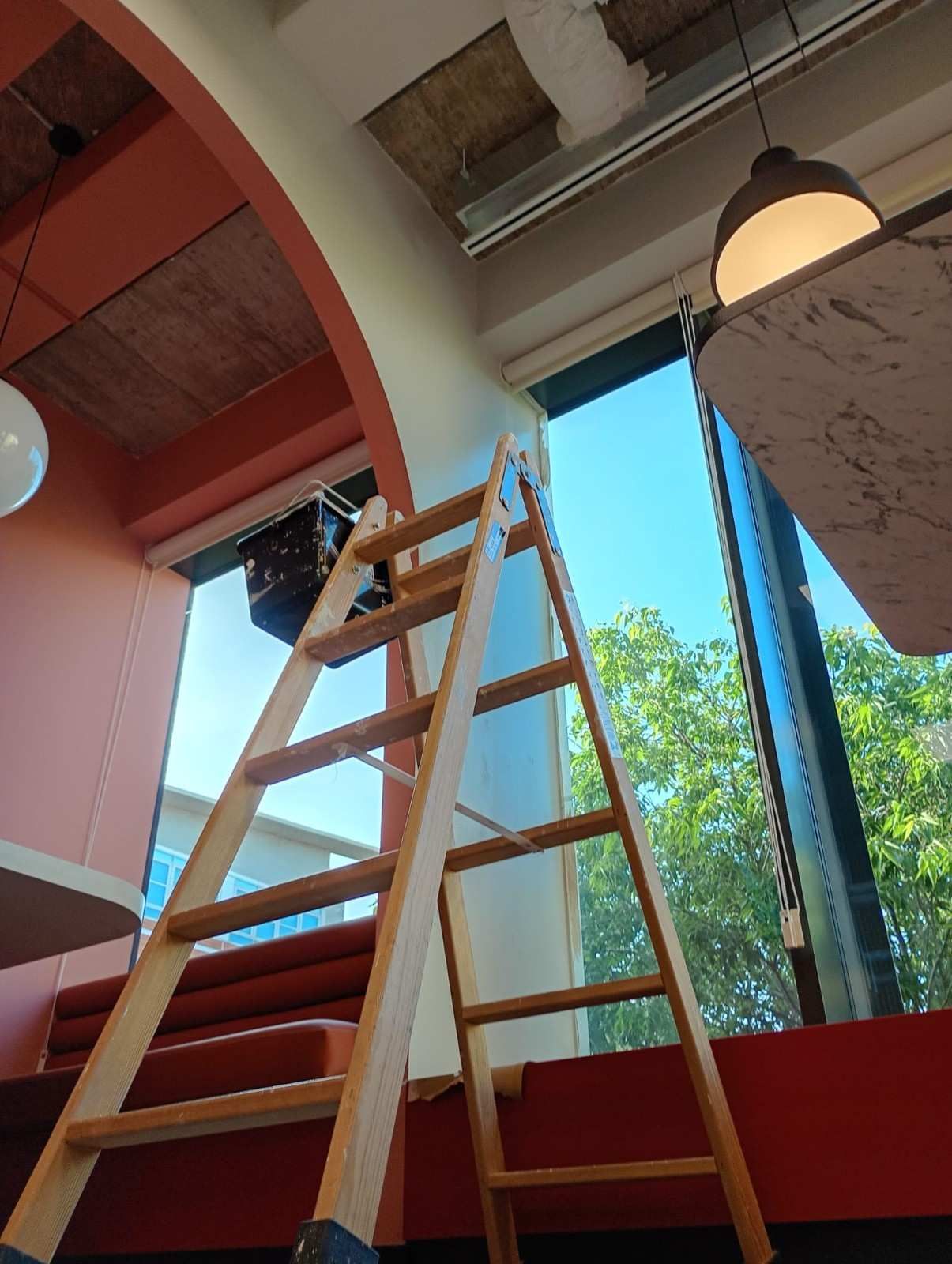 Escalera de madera apoyada contra una pared en un espacio interior luminoso con una gran ventana y luces colgantes.
