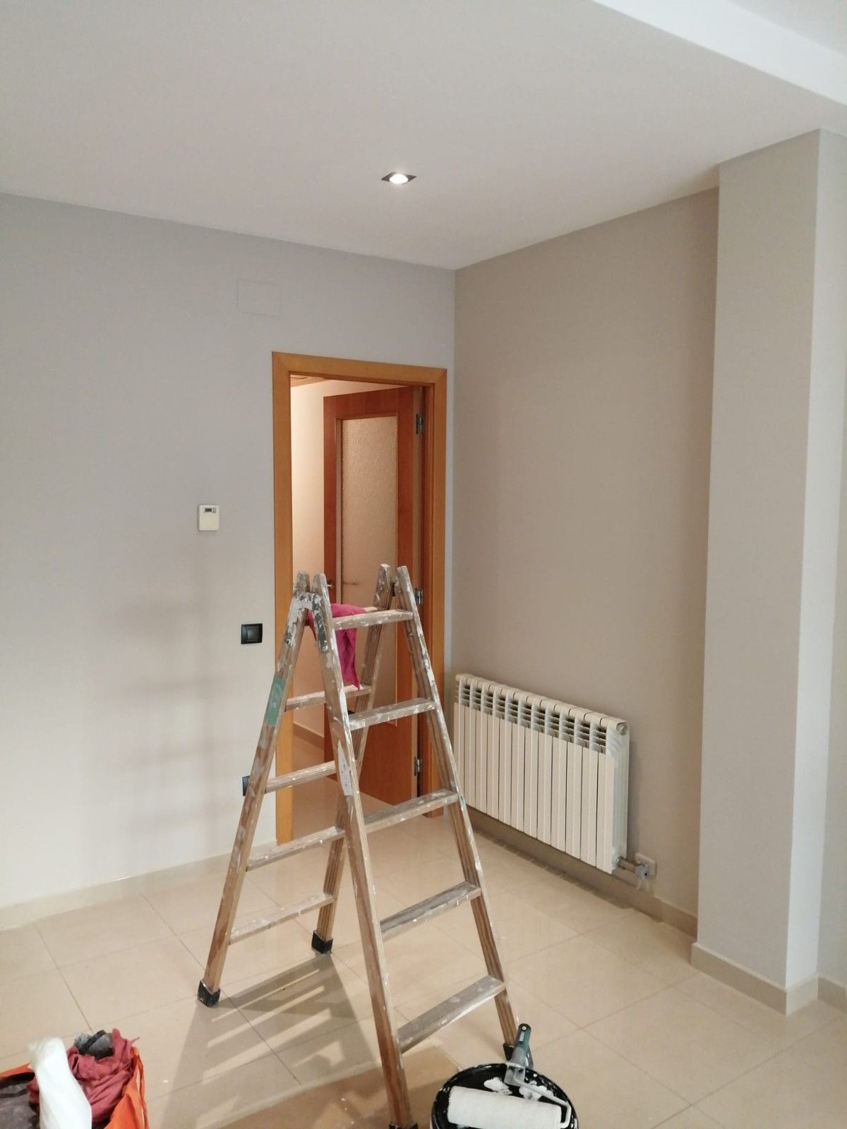 Una habitación vacía con una escalera en el centro, paredes de color beige y un radiador blanco cerca de una puerta abierta.