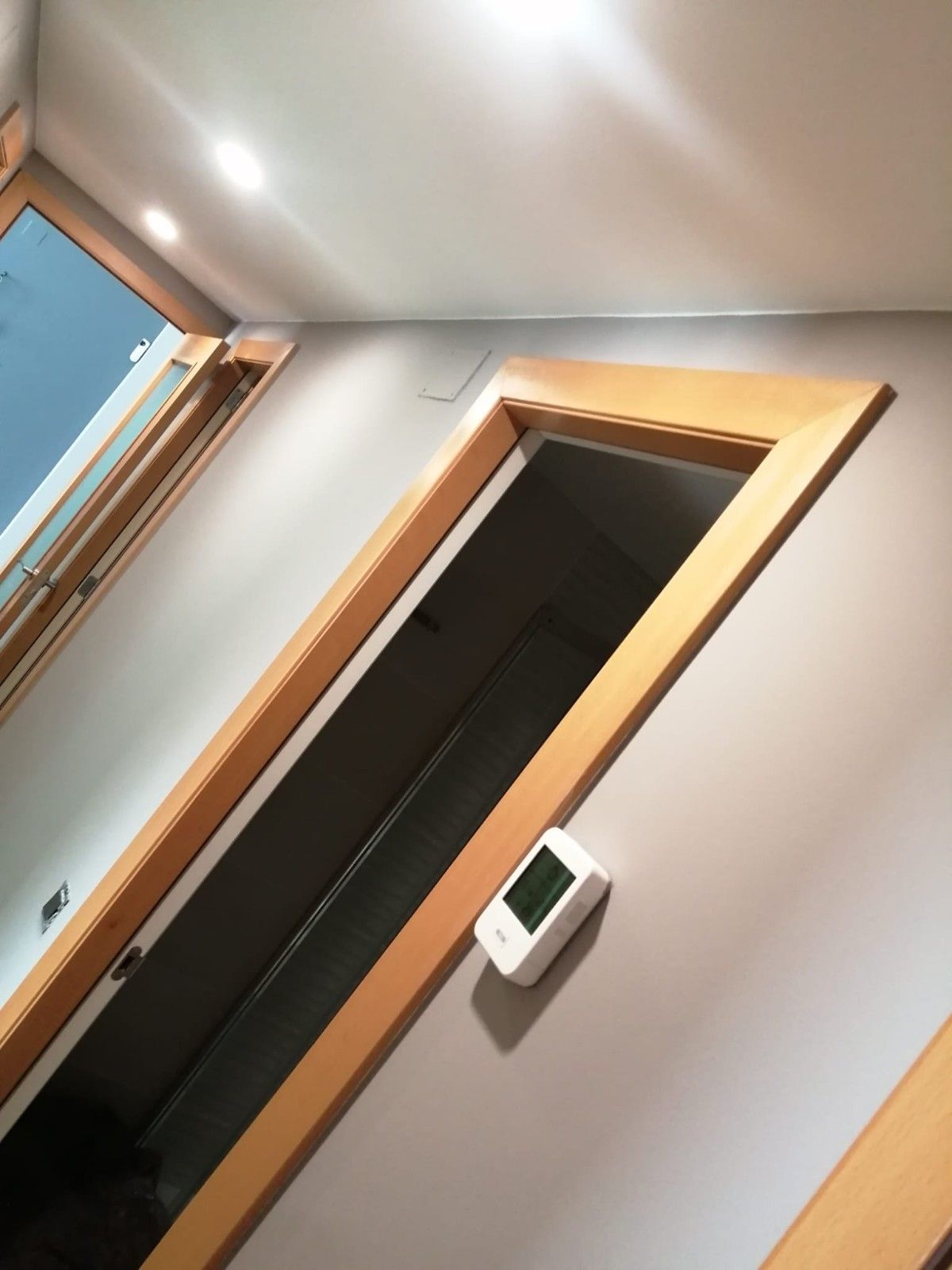 Vista inclinada de un pasillo con una puerta de madera, un termostato y un marco de ventana sobre paredes de color beige.