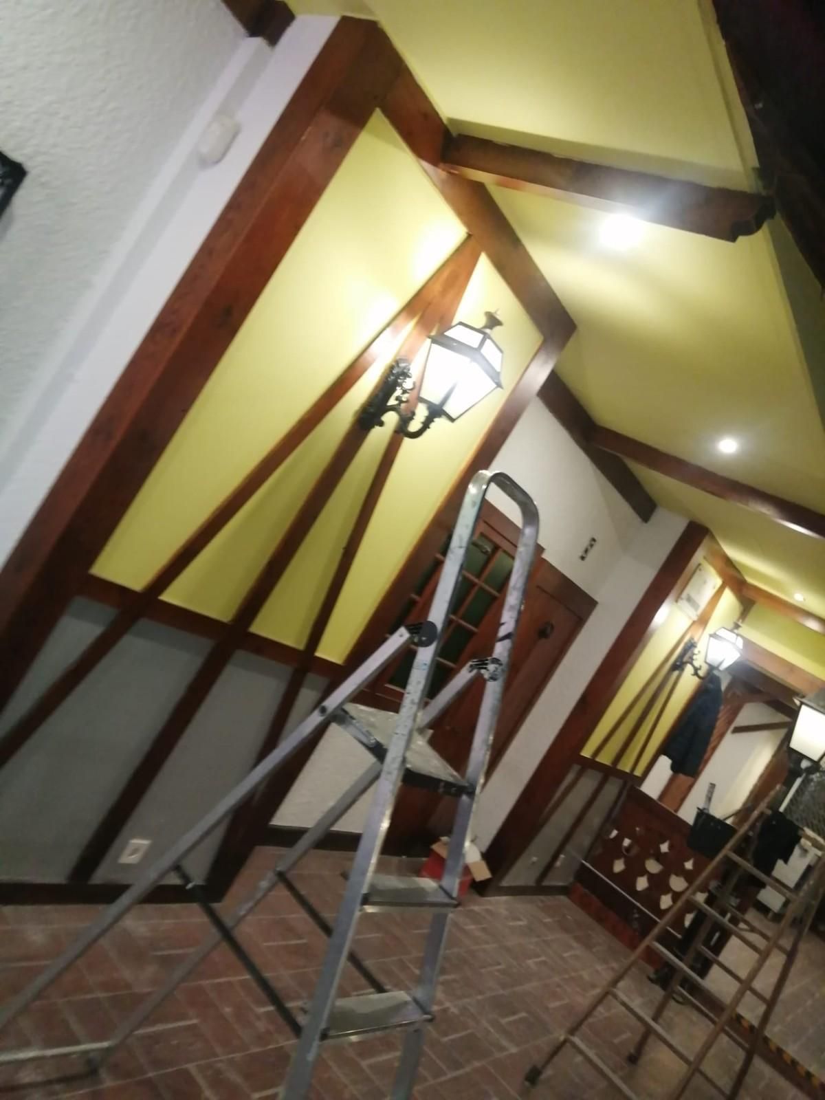 Pasillo inclinado con paredes amarillas, vigas de madera, una escalera y una lámpara de pared encendida.