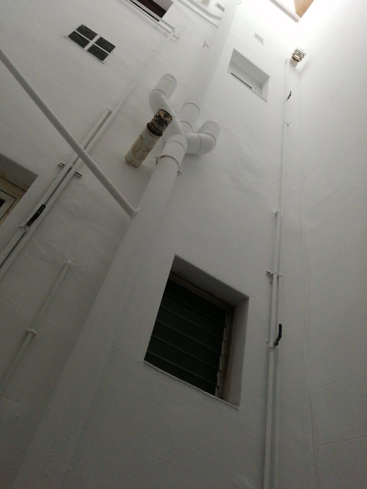 Patio de un edificio alto y blanco con ventanas, balcones y una tubería montada en la pared, visto desde abajo.