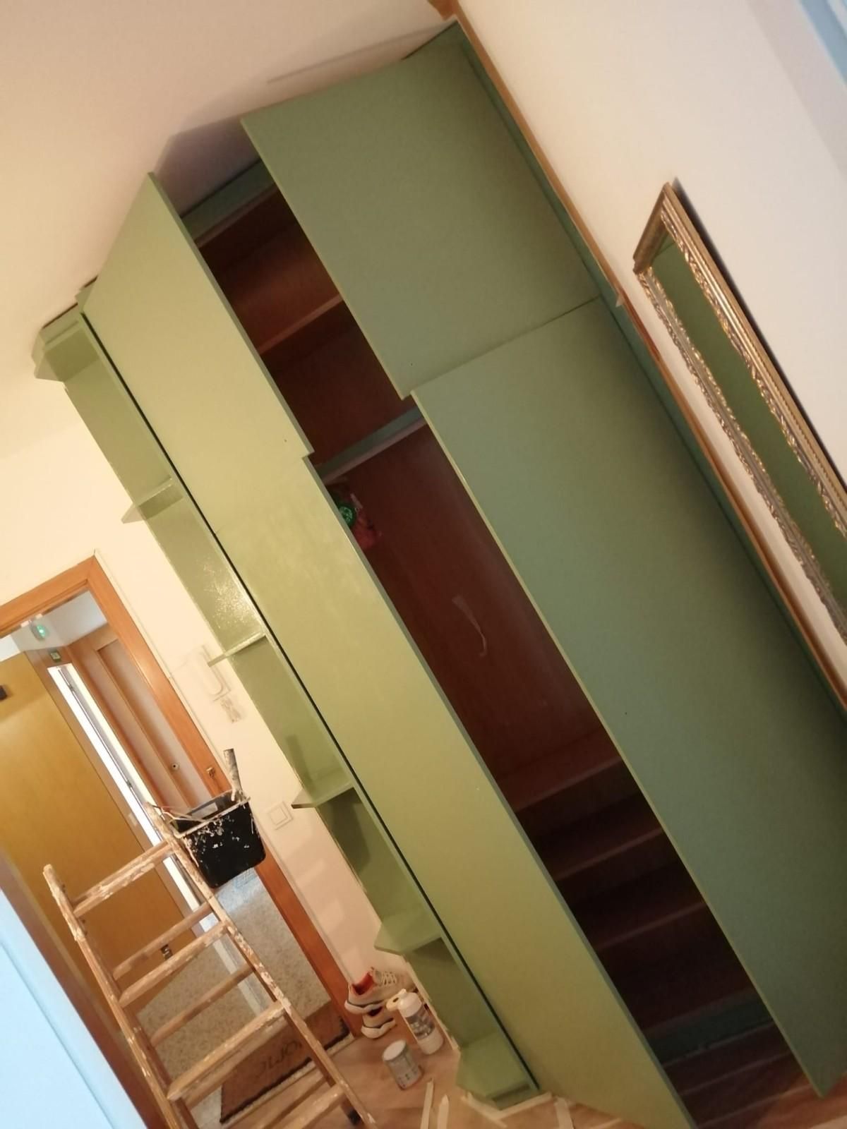 Vista inclinada de un pasillo con un armario alto verde, una escalera y un espejo de pared enmarcado.