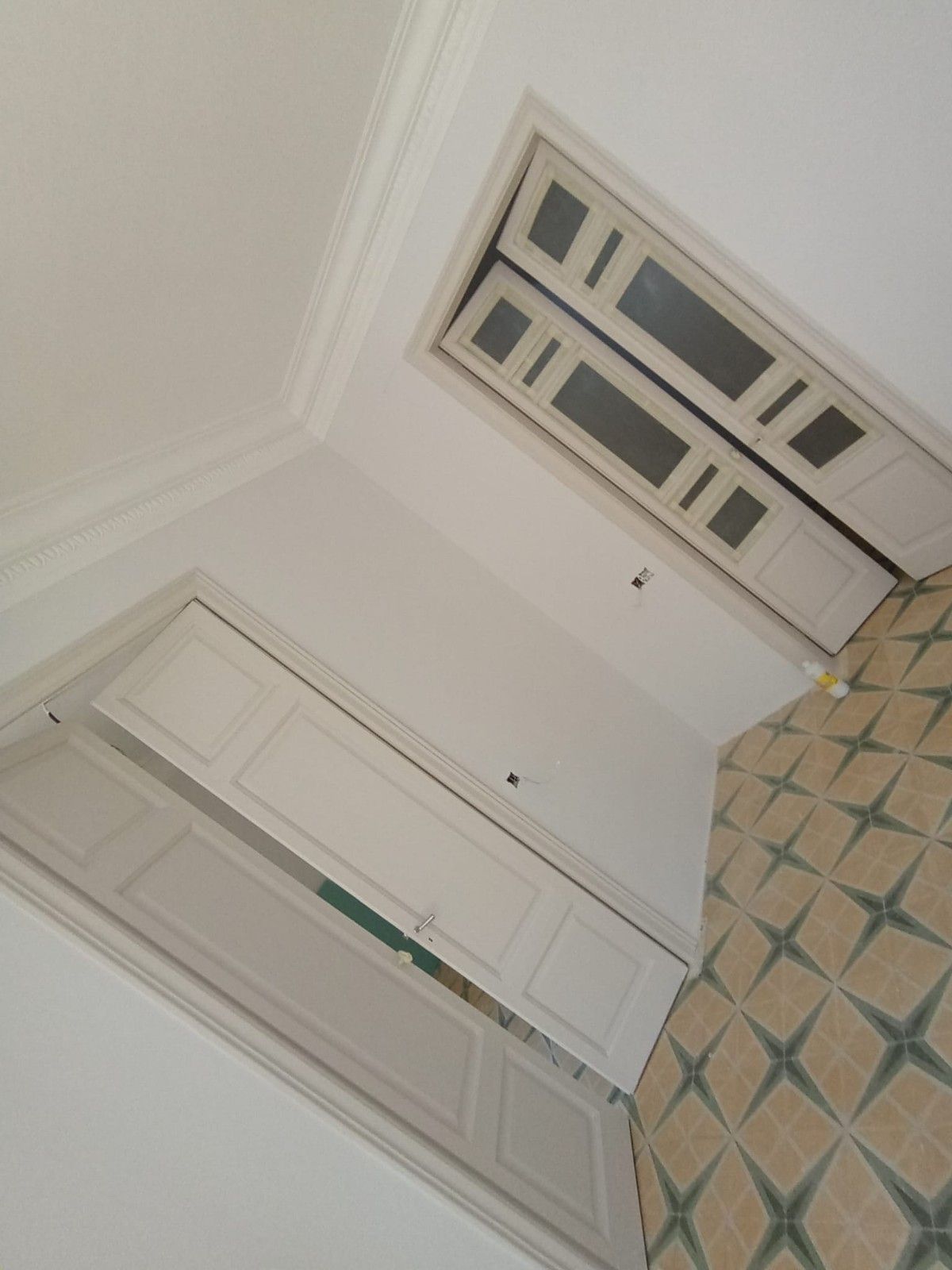 Vista inclinada de una habitación con paredes blancas, puertas dobles y una alfombra estampada cerca de una ventana.