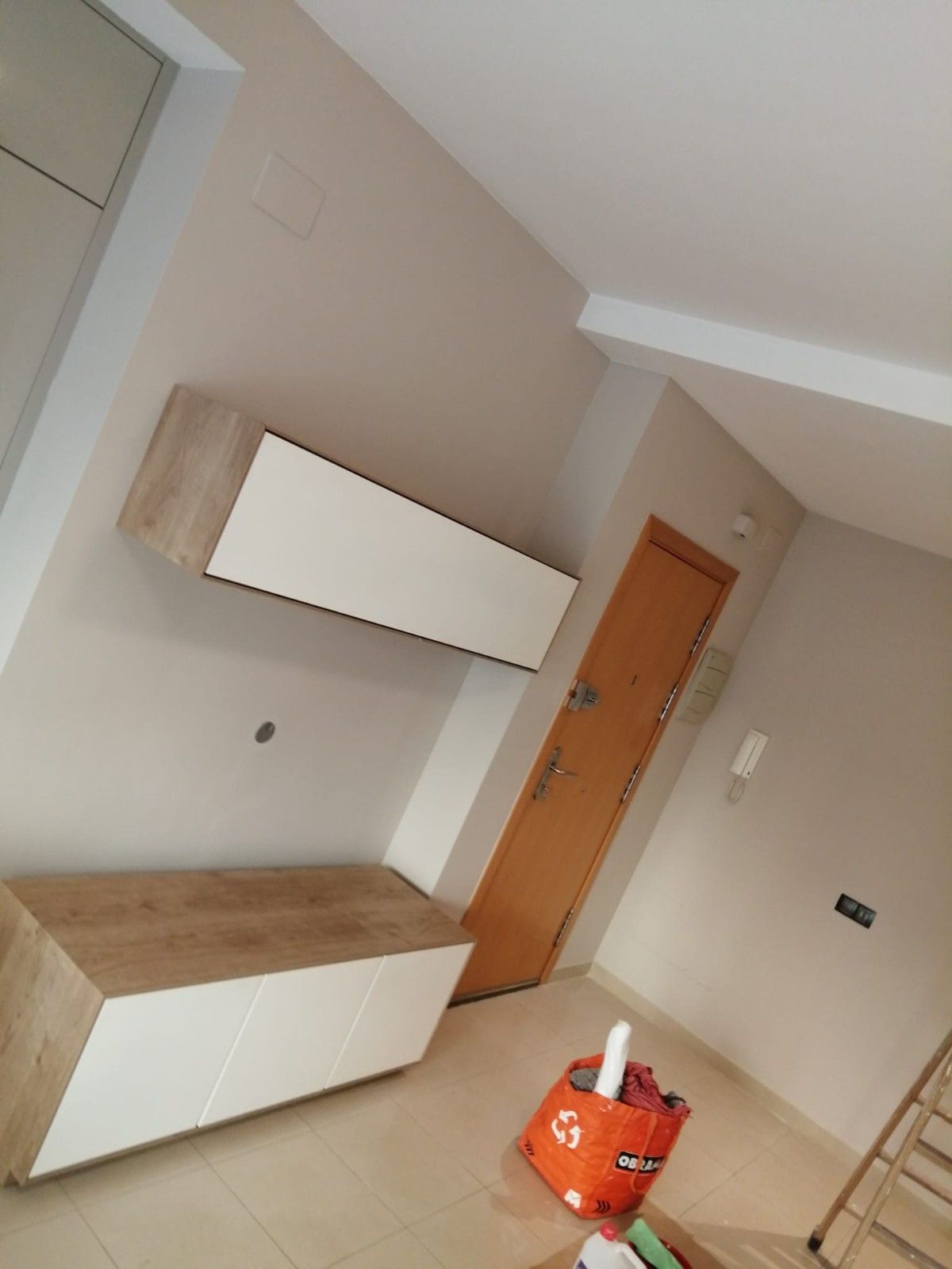 Habitación minimalista con paredes beige, armarios de madera y blancos, una puerta de madera y una bolsa naranja en el suelo.