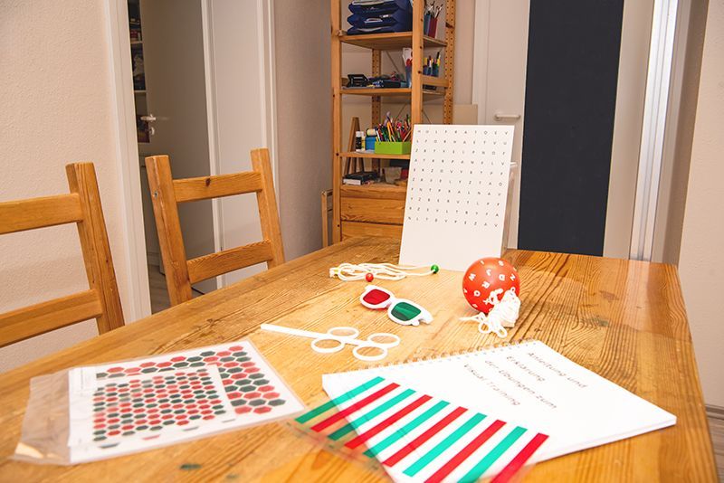 Holztisch mit Sehtestinstrumenten: Brille, Rot/Grün-Streifen, Punkttafel, Notizbuch und ein roter Ball.