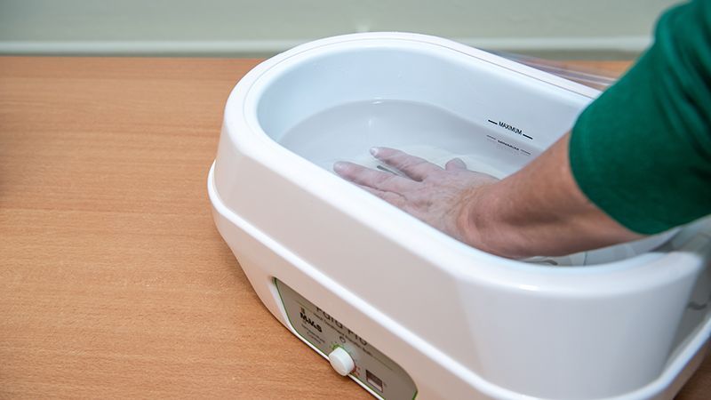 Hand in ein warmes Paraffinwachsbad getaucht, mit einem weißen Behälter auf einer Holzoberfläche.