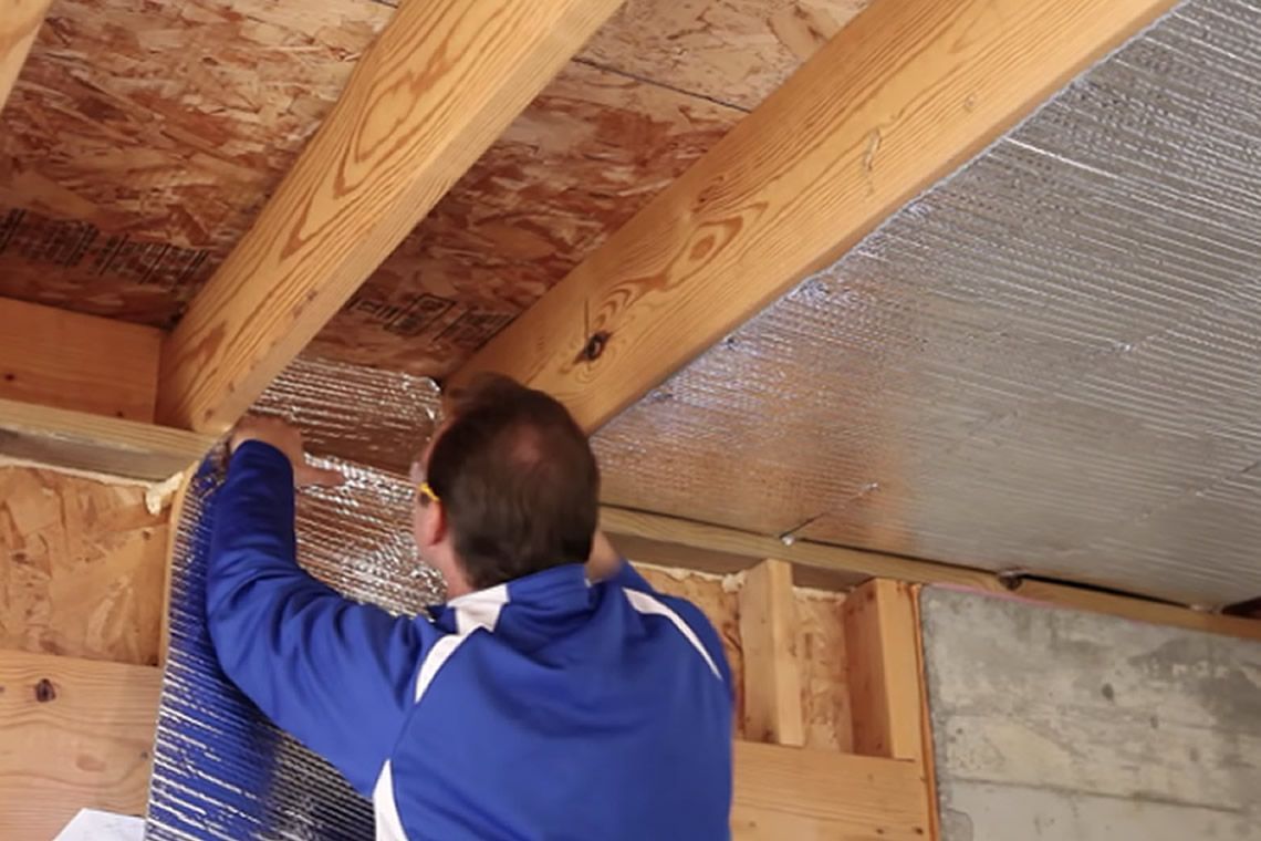 Hombre instalando aislamiento de aluminio en un techo con vigas de madera expuestas.