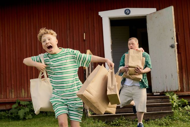 Etualalla nuori poika juoksee ulos käsityöläismökistä käsissään ostoskasseja