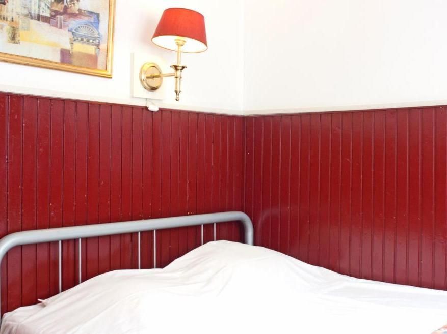 Red-paneled corner of a room with a bed covered in a white sheet. A wall lamp and a framed picture are visible.