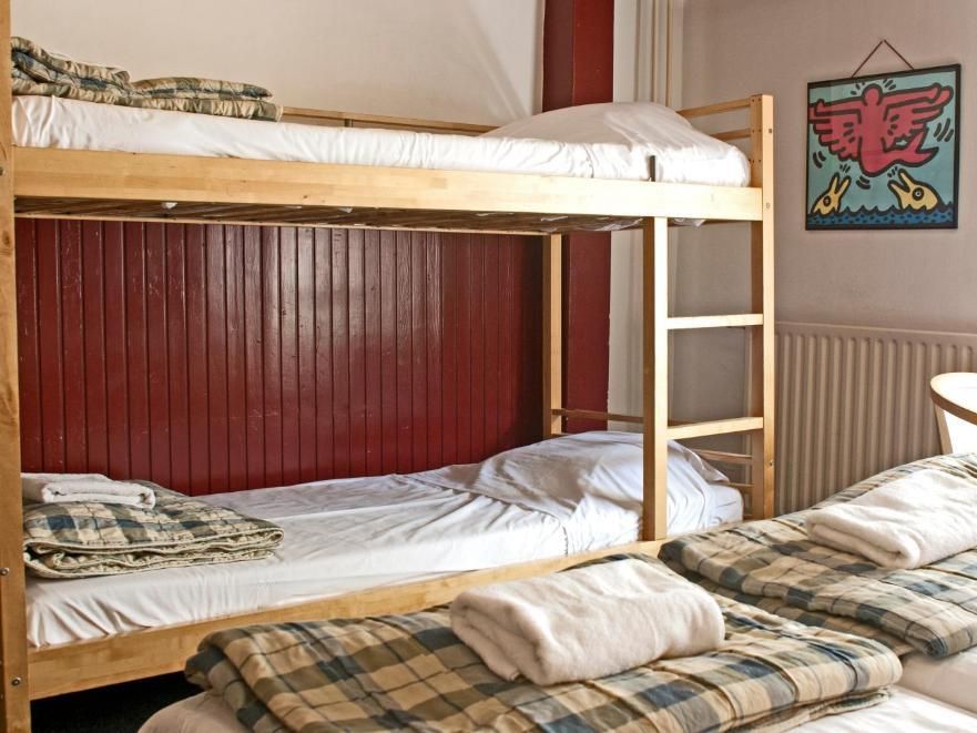 A hostel room with a bunk bed and two single beds, all made with white sheets and plaid blankets. A colorful artwork hangs on the wall.
