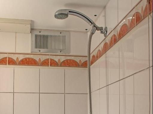 Showerhead and vent in a bathroom with white and orange-trimmed tiles. Chrome fixtures against a white tiled wall.
