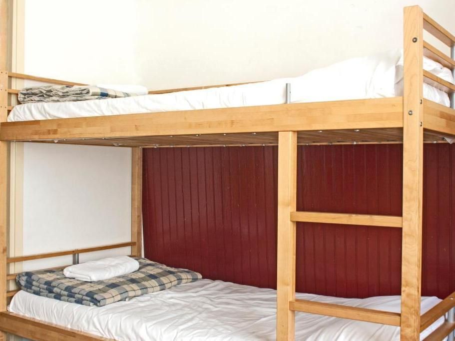 Bunk beds with wooden frame, white sheets, and a plaid blanket. Red paneling is behind the beds.