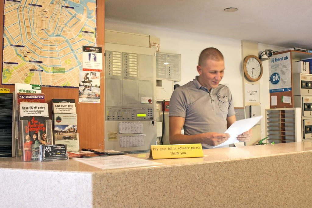 Een man bij de receptie van een hotel, glimlachend terwijl hij papieren bekijkt. Een kaart en brochures hangen aan de muur en de balie.